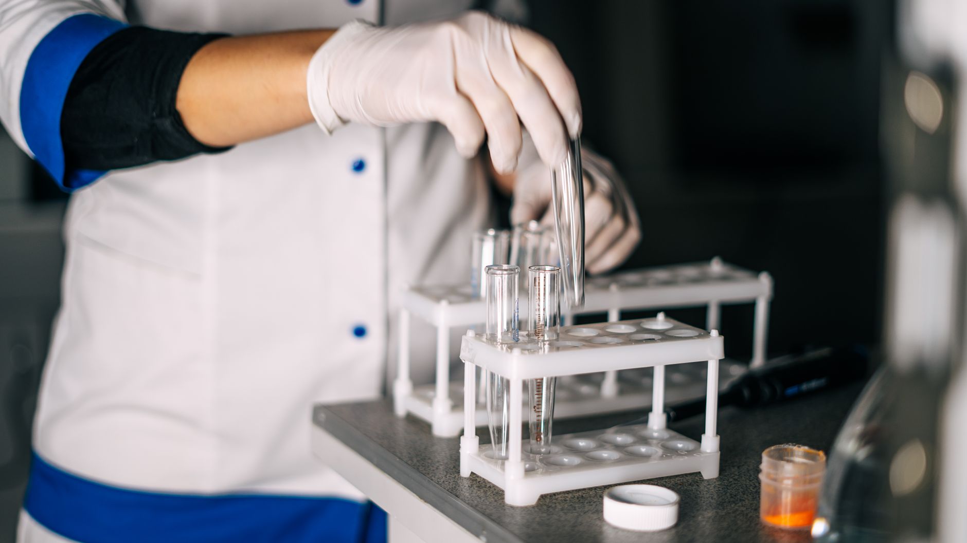 Gloved hands placing a glass test tube into a rack in a laboratory setting.