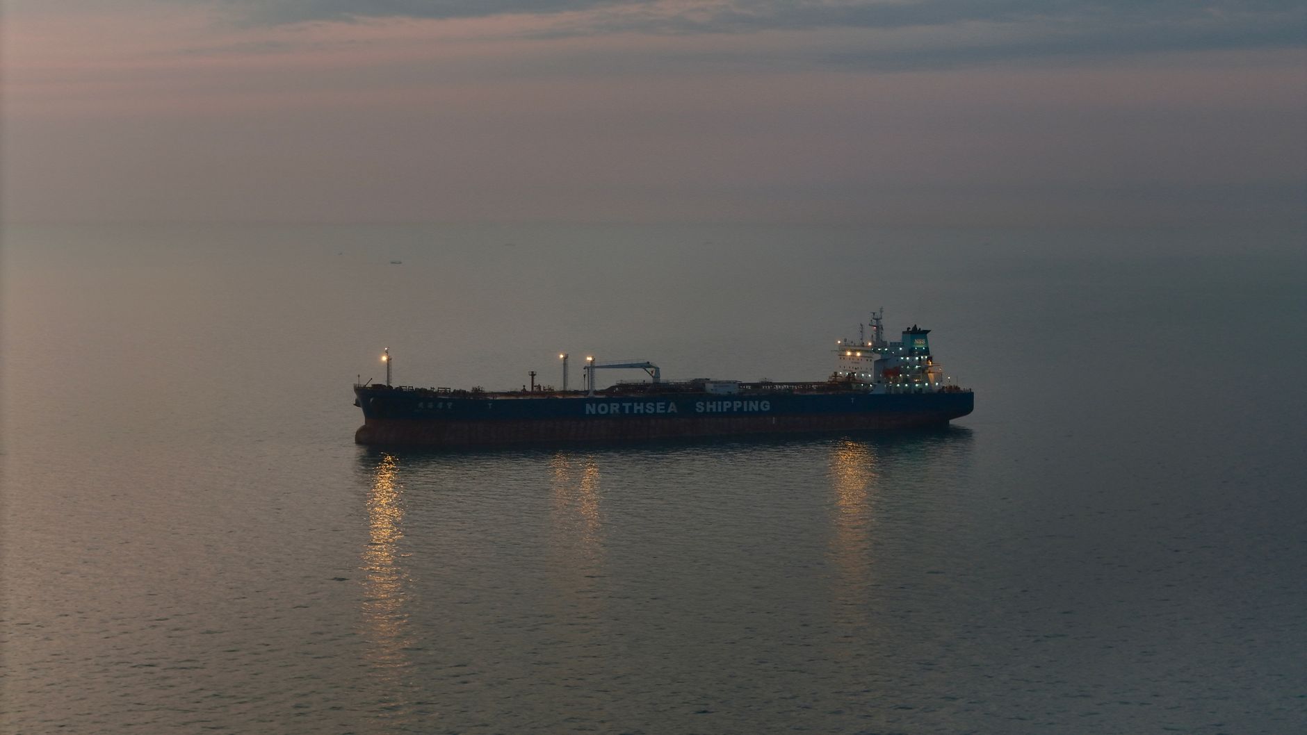 Oil tanker at sea at dusk, its deck lights reflected on calm water under a hazy sky.