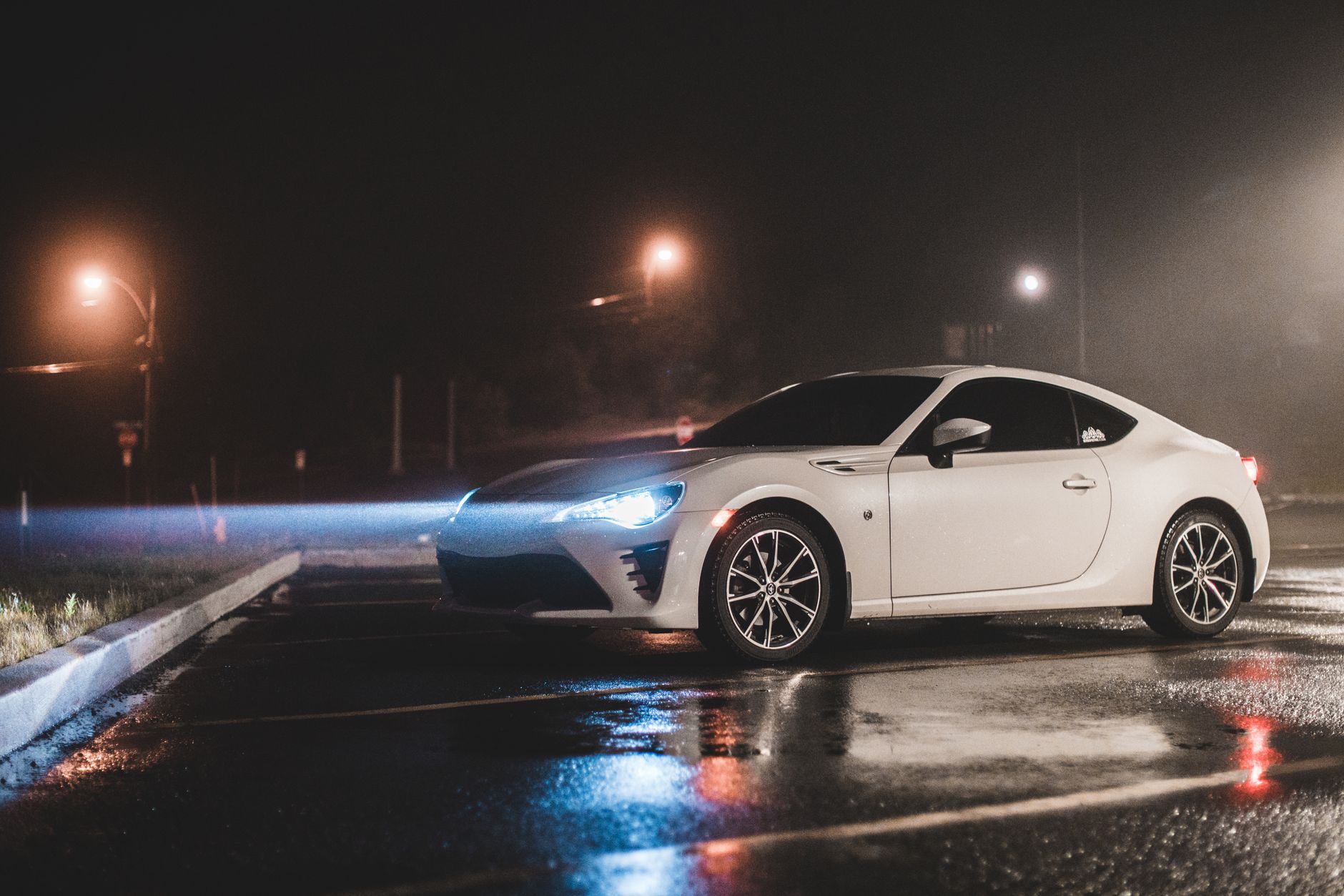 White coupe with headlights on, parked at night on wet asphalt under misty street lamps, with light reflections on the ground.