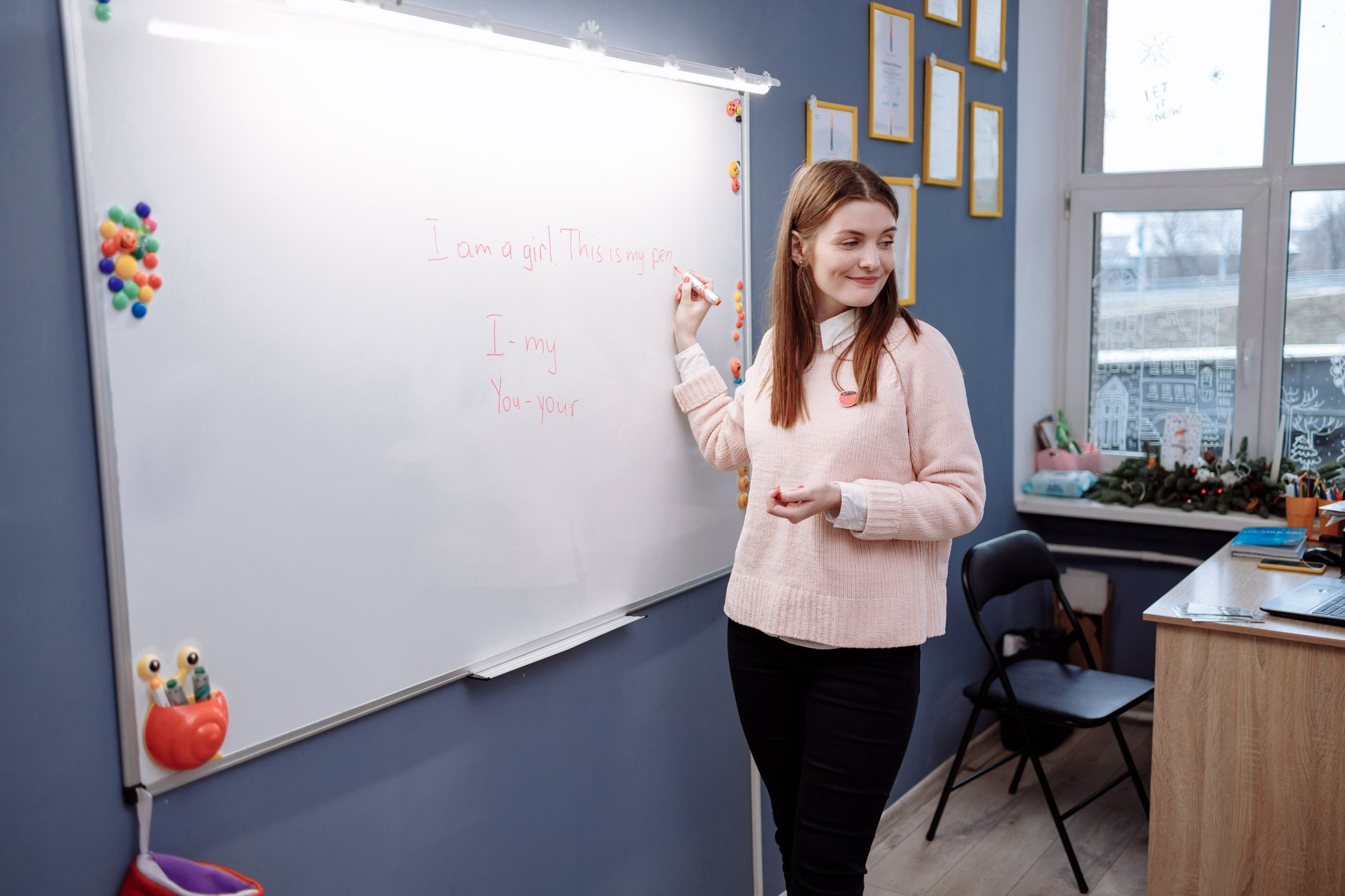 Woman in a pink sweater writes English phrases on a whiteboard in a classroom with a blue wall, a folding chair and a desk.